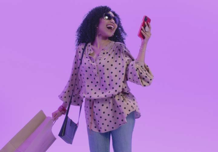 a smiling woman holding a shopping bag and looking at her phone in front of a purple background