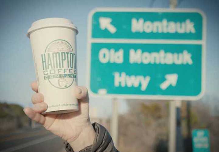 a womans hand holding a Hampton Coffee Company coffee cup in front of a green street sign pointing to Montauk