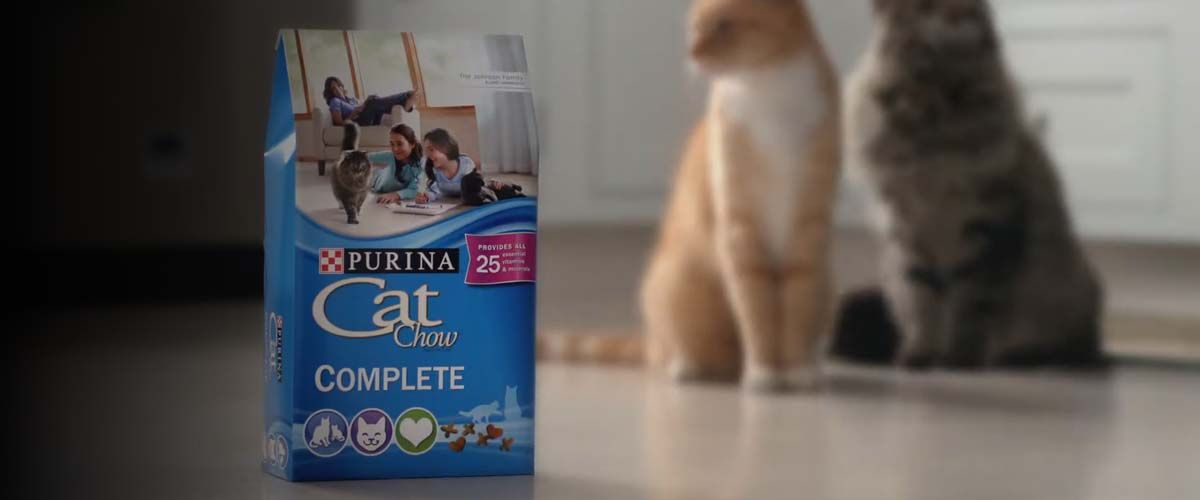 a Purina Cat Chow on a kitchen floor with two cats sitting patienly behind it
