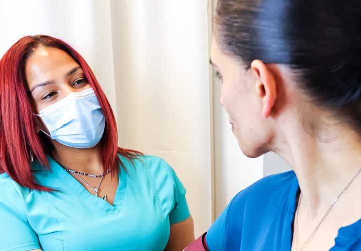 a smiling nurse in scrubs and mask talking to a female patient